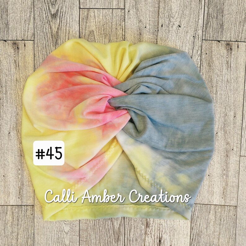 Op de afbeelding: Een tie-dye tulband met een gedraaid ontwerp aan de voorzijde in gemengde kleuren geel, roze en blauw. De stof heeft een zacht, gem&ecirc;leerd patroon. Een wit vierkant label aan de linkerkant bevat de tekst #45 en Calli Amber Creations.