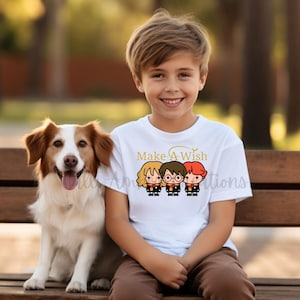 May include: A white t-shirt with a gold graphic of Harry Potter, Ron Weasley, and Hermione Granger characters from the Harry Potter series. The text "Make-A-Wish" is above the characters. A young boy is wearing the shirt and smiling.