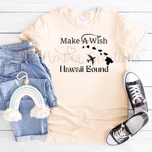 Puede incluir: Una camiseta beige con el texto "Make A Wish Hawaii Bound" y un avión volando sobre una cadena de islas. La camiseta está colocada sobre una superficie de madera con un par de pantalones vaqueros azules y una pared de macramé arcoíris.
