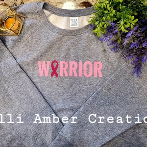May include: Gray sweatshirt with the word "WARRIOR" in pink block letters and a pink ribbon graphic. The sweatshirt has long sleeves and a crew neck. The text "Calli Amber Creations" is printed at the bottom.