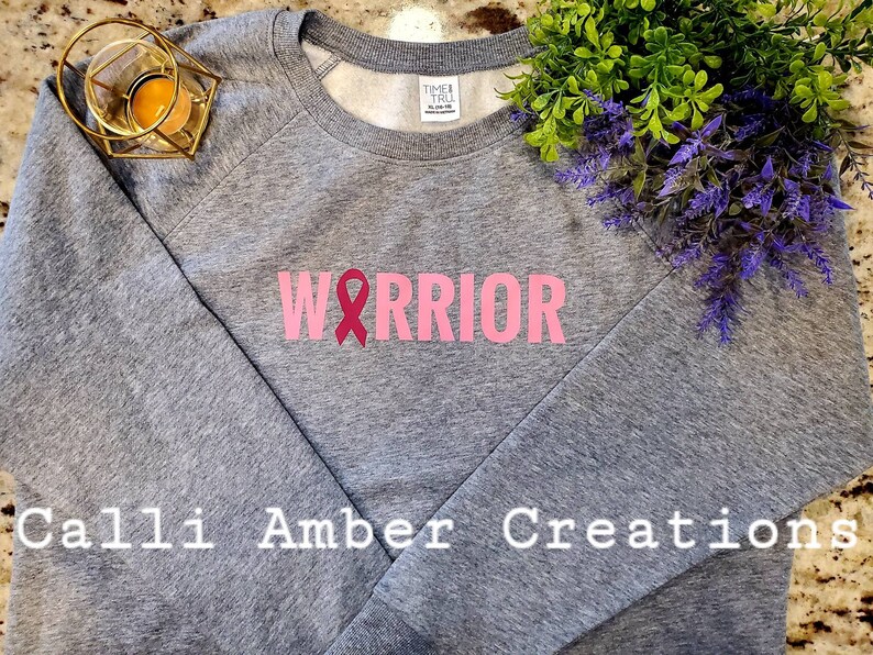 May include: Gray sweatshirt with the word "WARRIOR" in pink block letters and a pink ribbon graphic. The sweatshirt has long sleeves and a crew neck. The text "Calli Amber Creations" is printed at the bottom.