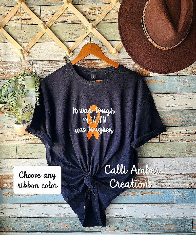 May include: Black t-shirt with the text "It was tough but CADEN was tougher" and an orange ribbon. The text "Calli Amber Creations" is also on the shirt. Choose any ribbon colour.