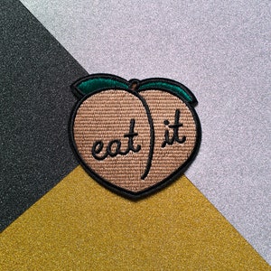 Peut inclure: Un patch brodé en forme de pêche avec le texte "eat it" en fil noir. Le patch est brun avec des coutures vertes autour des bords.