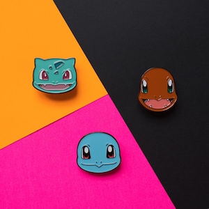 Peut inclure: Trois épingles en émail représentant les visages de dessin animé des personnages Pokémon Bulbizarre, Salamèche et Carapuce. Les épingles sont sur un fond coloré.