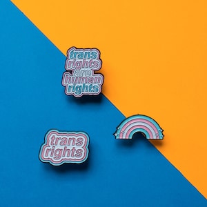 Może przedstawiać: Dwa szpilki emaliowane na niebieskim i żółtym tle. Jeden szpilka to tęcza z gwiazdami. Drugi szpilka głosi "trans rights are human rights" w różowych i niebieskich literach.