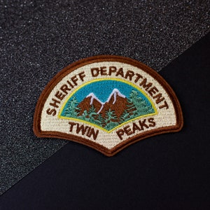 Peut inclure: Un écusson brodé marron et beige avec le texte "Sheriff Department Twin Peaks" en blanc. L'écusson représente une image stylisée d'une chaîne de montagnes avec des pins.