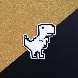 Peut inclure: Un écusson de dinosaure pixélisé blanc et noir. L'écusson présente un design rétro en 8 bits d'un Tyrannosaurus Rex. L'écusson est sur un fond or et noir.