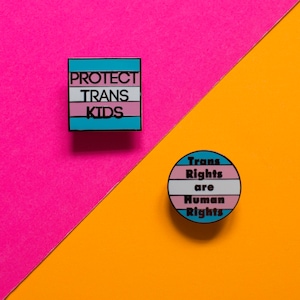 Könnte beinhalten: Zwei Anstecknadeln mit einem Transgender-Pride-Flaggen-Design. Die erste Nadel ist quadratisch und sagt "Protect Trans Kids". Die zweite Nadel ist rund und sagt "Trans Rights are Human Rights".