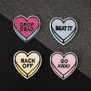 May include: Four heart-shaped patches with different sayings. The patches are pink, blue, yellow, and white with black text. The sayings are "Drop Dead", "Beat It", "Back Off", and "Go Away".