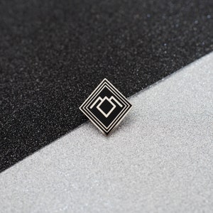May include: Black and white enamel pin with a geometric design. The pin features a square with a smaller square inside, and a series of lines forming a mountain-like shape.