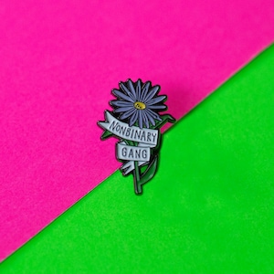 Könnte beinhalten: Eine schwarze Emaille-Anstecknadel mit einer lila Blume und einem weißen Banner mit der Aufschrift "Nonbinary Gang" auf einem rosa und grünen Hintergrund.