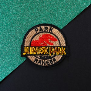 Peut inclure: Un écusson brodé marron et noir avec une silhouette de dinosaure rouge et le texte "Jurassic Park Ranger" en lettres jaunes et noires.