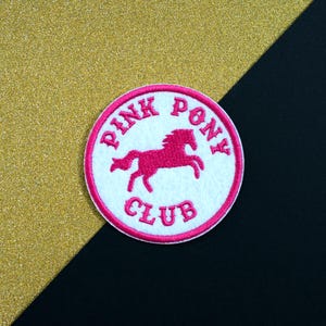 Op de afbeelding: Een ronde, witte patch met een roze rand en de woorden "PINK PONY CLUB" in roze letters. Een roze silhouet van een paard staat in het midden. De achtergrond is goud en zwart.