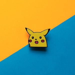 Peut inclure: Épingle en émail jaune représentant le visage surpris de Pikachu, un personnage de la franchise Pokémon. L'épingle a des contours noirs, des joues rouges et une expression surprise. Le fond est divisé en diagonale, avec des sections orange et bleu.