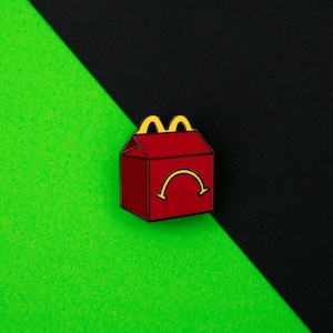 Peut inclure: Épinglette rouge en forme de boîte Happy Meal, avec un dessus en arc jaune et un visage triste. L'épinglette est posée sur un fond diagonal noir et vert.