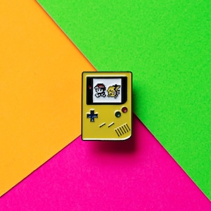 Op de afbeelding: Gele emaille pin in de vorm van een draagbare videogameconsole met een gepixelde afbeelding van een personage uit een populaire videogame-serie op het scherm.