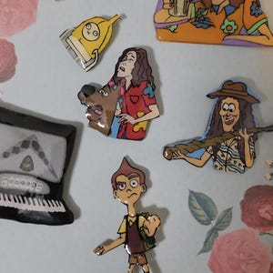 May include: A collection of colorful cartoon character pins. The pins feature various characters, including a person with glasses and the number 2025, a yellow object, a person with long hair holding a drink, and a person with a dog.