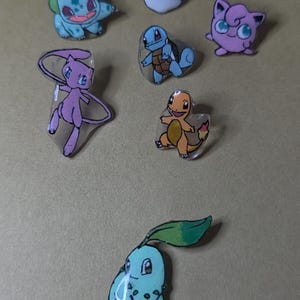 May include: A collection of colorful, cartoon-style pins featuring various Pokémon characters, including Bulbasaur, Squirtle, Charmander, Jigglypuff, Mew, and a Poké Ball. The pins are set against a neutral background.