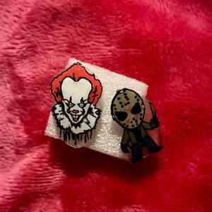May include: Two horror-themed rings. One ring features a cartoon image of Pennywise the clown with red hair and a white ruffled collar. The other ring depicts Jason Voorhees with a hockey mask and a knife. Both rings are on a white surface.