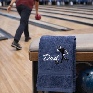 May include: A navy blue terry cloth towel featuring white embroidered text that reads Dad, accompanied by an embroidered graphic of a person in a black shirt throwing a bowling ball, with a textured border detail near the bottom edge.