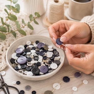 May include: A collection of round sewing buttons in shades of white, black, grey, and purple contained within a speckled ceramic bowl. Some buttons are threaded onto a piece of twine, while others remain loose in the bowl alongside metal scissors.