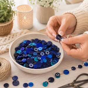 May include: A collection of round buttons in various shades of blue, including navy, royal, and turquoise, contained within a speckled ceramic bowl. Some buttons feature white circular patterns, and several are threaded onto a piece of twine.