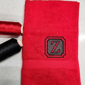 May include: A folded, bright red bath towel featuring an embroidered black and grey letter "Z" within a decorative frame. Two thread spools, one red and one black, are placed near the towel. The towel has a textured surface.