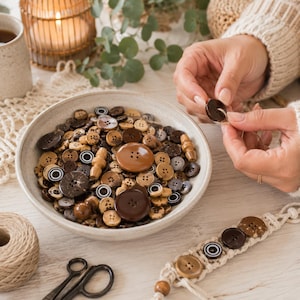 May include: A collection of assorted round buttons in various shades of brown, tan, and black, featuring different sizes and textures. The buttons are contained within a shallow, light-coloured ceramic bowl, with some buttons attached to a macrame cord.