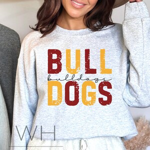 Puede incluir: Una sudadera gris con las palabras "BULLDOGS" en letras rojas y amarillas con un aspecto desgastado. El texto "WH WHITE HOUSE CO" est&aacute; en la parte inferior de la sudadera.