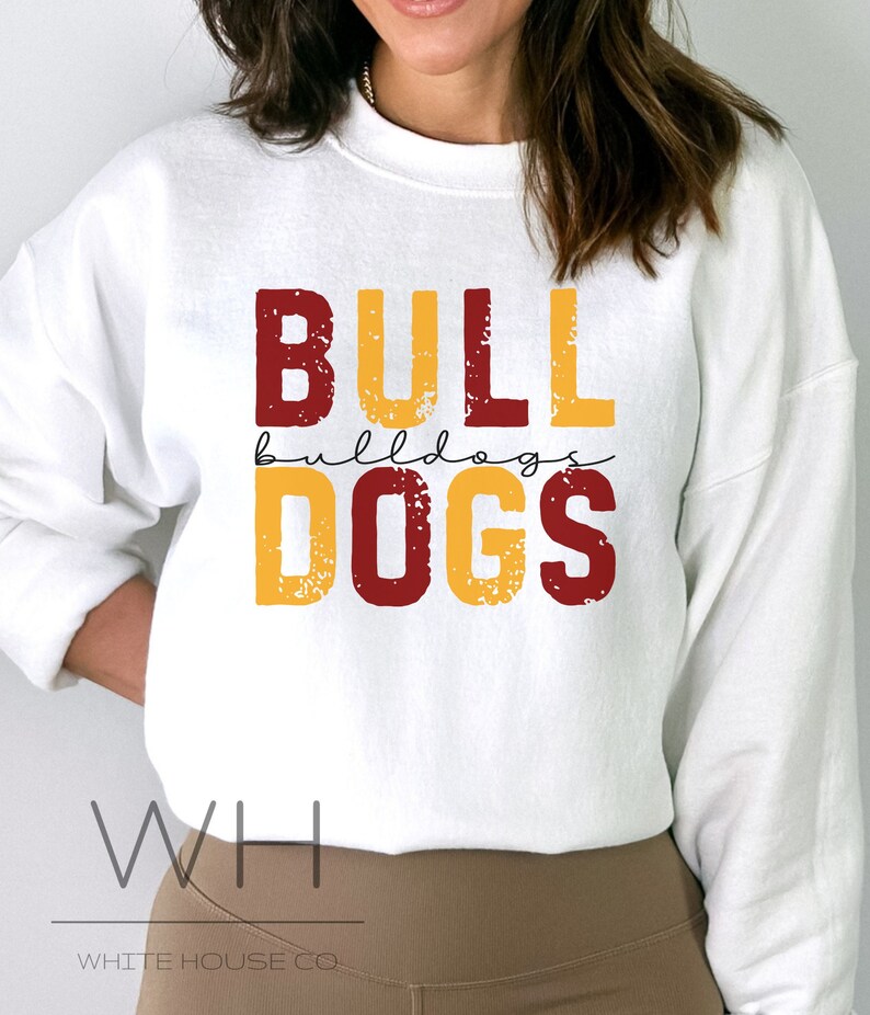 Puede incluir: Sudadera blanca con un gr&aacute;fico rojo y amarillo que dice "BULLDOGS" con la palabra "bulldogs" escrita en letras m&aacute;s peque&ntilde;as debajo de la palabra "BULL". El texto tiene un aspecto desgastado. La sudadera tiene las palabras "WHITE HOUSE CO." impresas en la parte inferior.