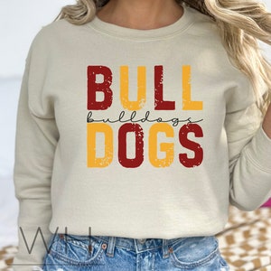 Puede incluir: Una sudadera beige con las palabras "BULLDOGS" impresas en letras rojas y amarillas con un aspecto desgastado. La palabra "bulldogs" est&aacute; escrita en cursiva en letras m&aacute;s peque&ntilde;as debajo de la palabra "BULL".