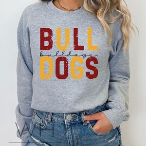 Puede incluir: Sudadera gris con un dise&ntilde;o gr&aacute;fico desgastado que dice "BULLDOGS" en letras rojas y amarillas.