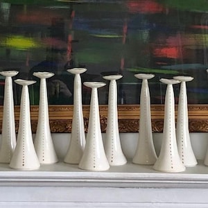 Op de afbeelding: Een verzameling van 14 witte keramische kaarsenstandaards met een gestippeld patroon. De kaarsenstandaards zijn in een rij op een wit oppervlak gerangschikt.