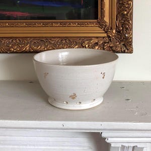 May include: White ceramic colander with gold speckles. The colander has a round shape and a flat bottom. It has small holes around the sides.