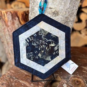 May include: Hexagonal quilted pot holder featuring a moose design. It has a dark blue border, a white and light blue geometric pattern, and a patterned hanging loop. The pot holder is displayed on a black metal stand. The tag reads "Handmade in Bush Alaska".