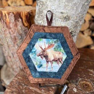 May include: Hexagonal quilted pot holder with a moose illustration. It has a brown and blue patterned border and a brown loop for hanging. The moose is shown in a naturalistic setting with water and trees.