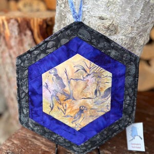 May include: Hexagon-shaped quilted wall hanging with a bird scene. The artwork features a central panel with a bird and landscape design, framed by a blue border and a patterned gray edge. A blue fabric loop is attached for hanging.