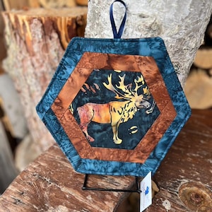 May include: Hexagonal quilted pot holder with a moose design. The pot holder has a teal, brown, and black colour scheme. A dark blue ribbon is attached for hanging. The moose is depicted in shades of yellow, orange, and brown.