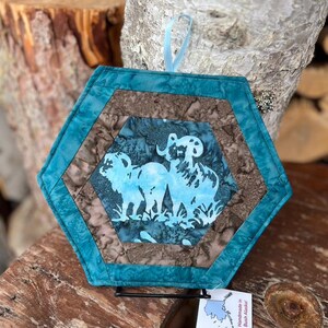 May include: Hexagon-shaped quilted potholder with a teal border and a brown and blue patterned center. The center features a light blue silhouette of two rams. A light blue ribbon is attached for hanging. The tag reads "Handmade in Bush Alaska!"