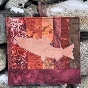 Könnte beinhalten: Ein quadratischer, gesteppter Topflappen mit einem Lachsmotiv. Der Topflappen hat ein Patchwork-Design in Rot-, Braun- und Burgundertönen. Eine Lachssilhouette ist auf der Vorderseite appliziert. Eine braune Schlaufe zum Aufhängen ist angebracht.
