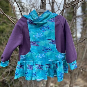 Könnte beinhalten: Eine lila-türkisfarbene Kinderjacke mit einem einzigartigen Design. Die Jacke hat eine türkisfarbene Front mit einem sich wiederholenden Flugzeugmuster, einen Rüschensaum und lila Ärmel. Die Kapuze und die Manschetten sind ebenfalls türkis. Die Jacke hängt an einem Kleiderbügel.