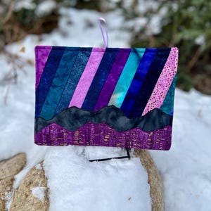 May include: A rectangular, quilted fabric item with a purple hanging loop. The design features diagonal stripes in shades of purple, blue, and teal, with a mountain range applique at the bottom. The item is resting on a snow-covered rock.