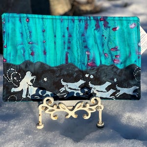 May include: A teal blue and black fabric coaster with a white silhouette of a dog sledding team. The coaster is on a white stand.