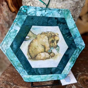 Pode incluir: Pega-panela hexagonal acolchoada com uma imagem em aquarela de um urso pardo segurando um peixe. Possui uma borda estampada em azul e verde-azulado e uma alça verde para pendurar. O urso está em um fundo branco.