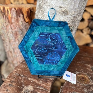 May include: Hexagon-shaped quilted potholder in shades of blue and teal. The center features a batik design with a floral pattern. A loop is attached for hanging. The potholder is displayed on a wooden surface.