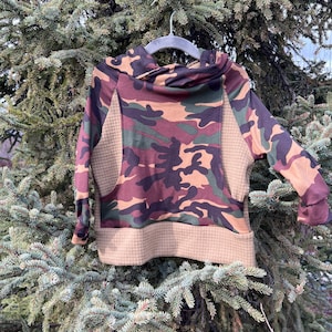 Op de afbeelding: Een camouflage-patroon hoodie met een col en lange mouwen. De stof heeft een mix van bruine, groene en bruine kleuren. De hoodie hangt aan een grijze hanger tegen een achtergrond van groen gebladerte.