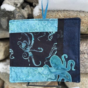 May include: Square quilted potholder with a blue ribbon loop. The potholder features a black panel with teal octopus designs, bordered by teal and navy blue fabric. The design is displayed on a black metal stand.