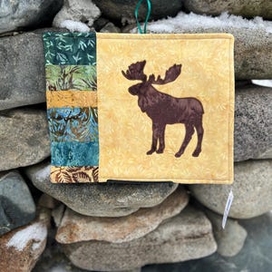 May include: A square, quilted pot holder with a moose applique. The pot holder features a yellow background with a brown moose silhouette. The left side has a patchwork design in shades of green, blue, and gold. A green ribbon loop is attached for hanging.