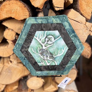 May include: Hexagonal quilted wall hanging with a nature-inspired design. The center features a bird perched on a branch with a full moon. The piece is framed with teal and black fabric and has a black hanging loop.