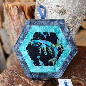 May include: Hexagon-shaped quilted pot holder with a whale design. The center features a black fabric with a blue and teal whale illustration. The pot holder is framed with teal and gray patterned fabric and has a blue hanging loop.